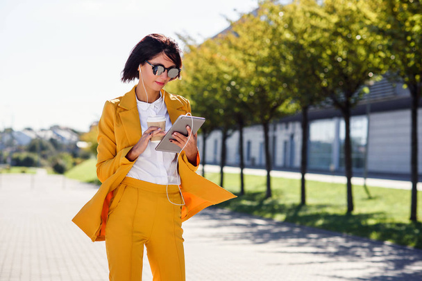 Stylish young business woman uses tablet computer while walks with cup of coffee on alley background. Attractive woman scrolling pages, tapping on touch screen, checking news and financial charts. - Foto, Imagem
