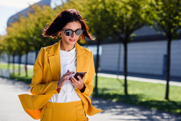 Stylish young business woman uses smart phone while walks near business centre on alley background. Attractive woman in suit scrolls pages, tapping on touch screen, checks news and financial charts. - Fotoğraf, Görsel