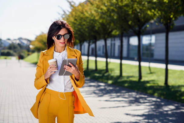 Stylish young business woman uses tablet computer while walks with cup of coffee on alley background. Attractive woman scrolling pages, tapping on touch screen, checking news and financial charts. - Photo, Image