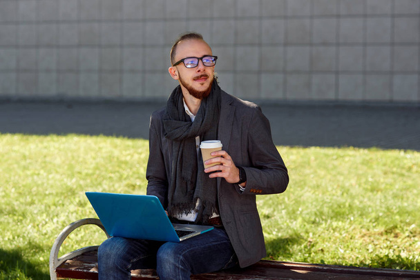 Young businessman in glasses dressed in jacket with scarf drinks coffee and using laptop on a lunch break outdoors sitting on the wooden bench in the park near business centre. - Foto, immagini