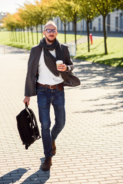 Молодой кавказский бизнесмен с рюкзаком и чашкой кофе на открытом воздухе возле современного бизнес-центра. Молодой бородатый бизнесмен, гуляющий по улице, пьет кофе во время обеденного перерыва
. - Фото, изображение