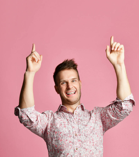 Young happy smiling man in shirt pointing fingers hands up on pink  - Photo, image