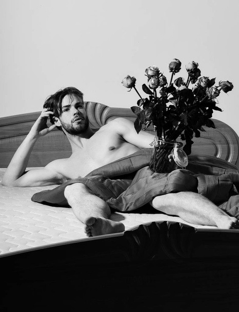young handsome bearded sexy macho man with stylish beard lying naked on bed under red blanket holding transparent vase with bouquet of orange roses and shows abs - Фото, изображение