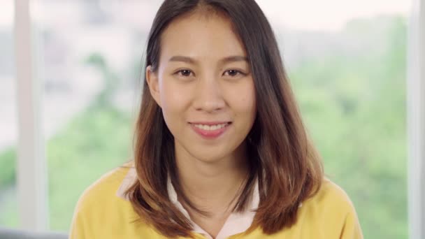 Zeitlupe - jugendliche asiatische Frau, die glücklich lächelnd in die Kamera blickt, während sie sich zu Hause in ihrem Wohnzimmer entspannt. schöne asiatische junge Frau mit entspannter Zeit zu Hause Konzept. - Filmmaterial, Video