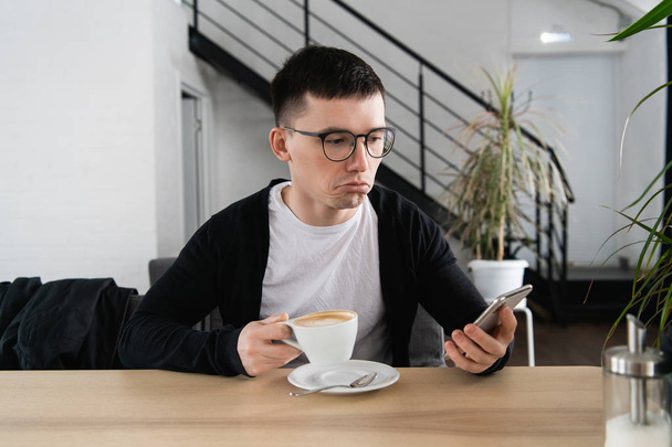 Worried young man reading a message with bad notification on modern smartphone sitting in cafe. Frustrated and upset hipster guy watching shocking video in blog on cellular - Fotó, kép