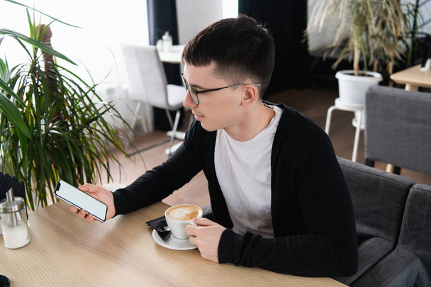 A photo of a handsome man wearing glasses looking at the screen of a mobile phone. Guy is sitting at the table of the coffee shop, holding smart-phone and relaxing. - Fotó, kép