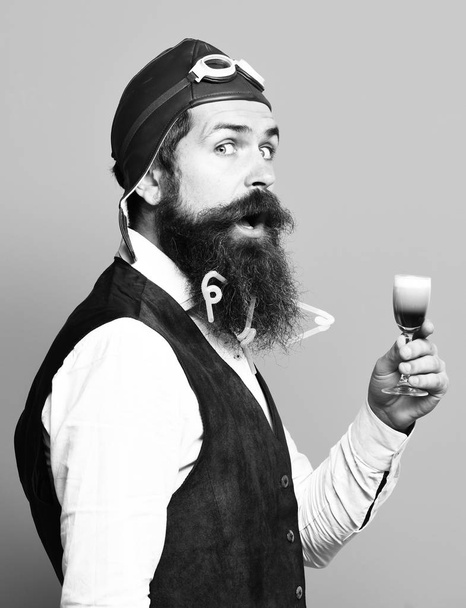 handsome bearded pilot or aviator man with long beard and mustache on funny face holding glass of alcoholic shot in vintage suede leather waistcoat with hat and glasses on blue studio background - Zdjęcie, obraz