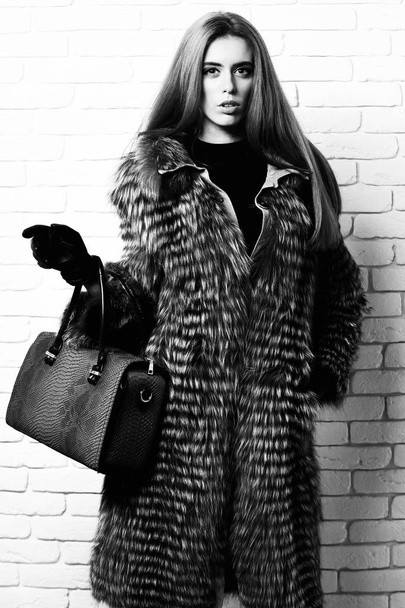 young fashionable sexy pretty rich woman with beautiful long blonde hair in waist coat of grey fur with black gloves showing blue leather bag with snake print on brick wall studio background - Foto, Imagem