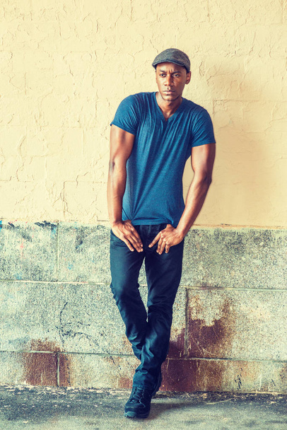 Retrato do jovem afro-americano em Nova York, vestindo azul V pescoço T-shirt, boné plano, preto sneaks, endireitando os braços, ombros encolhendo, de pé por parede pintada na rua, olhando, pensando
 - Foto, Imagem
