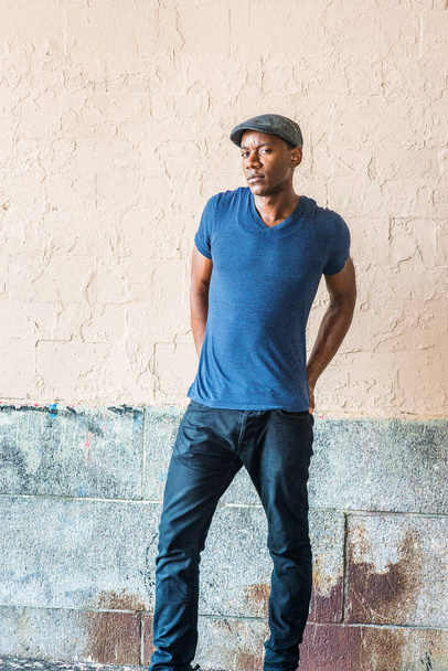 Young African American Man in New York, wearing blue V neck T shirt, flat cap, black pants, shrugging shoulders, arms backward down, hands on back, standing by painted wall on street, frowned, looking - Photo, Image