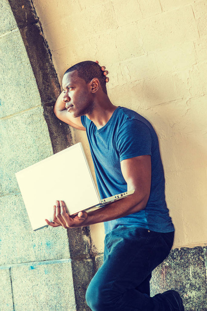 Молодой афроамериканец, работающий на ноутбуке в Нью-Йорке, одет в синюю футболку V-образной шеи, стоит напротив стены на улице, поднимает руку к стене, руки на затылке, смотрит в сторону
 - Фото, изображение
