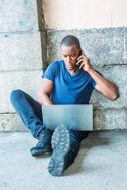 Joven afroamericano viajando, trabajando en Nueva York, vistiendo camiseta azul con cuello en V, pantalones negros, zapatillas de deporte, sentado en el suelo por la pared en la calle, trabajando en el ordenador portátil, hablando por teléfono celular
 - Foto, Imagen