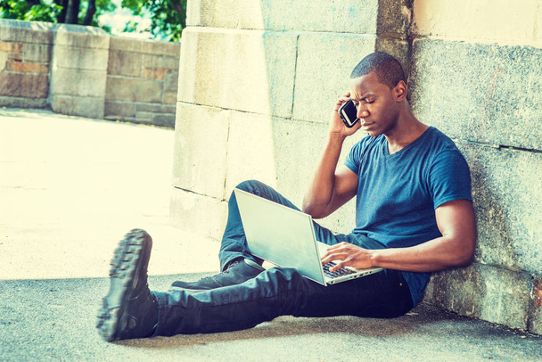 Young African American Man traveling, working in New York, wearing blue V neck T shirt, black pants, sneakers, sitting on ground by wall on street, working on laptop computer, talking on cell phone - Photo, Image