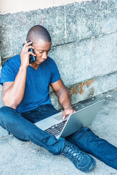 Young African American Man traveling, working in New York, wearing blue V neck T shirt, black pants, sneakers, sitting on ground by wall on street, working on laptop computer, talking on cell phone - Foto, immagini
