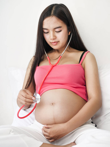 Aasialainen raskaana oleva nainen, joka käyttää punaista stetoskooppia kuunnellen vauvaansa sängyssä, elämäntapa käsite
. - Valokuva, kuva