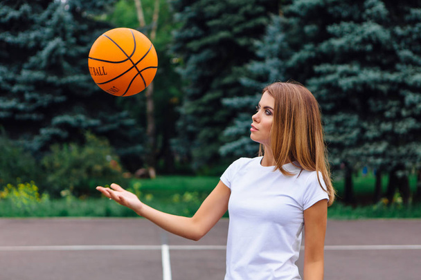 Beautiful young girl dressed in white t-shirt, shorts and sneakers, plays with a ball on a basketball court. - Foto, imagen
