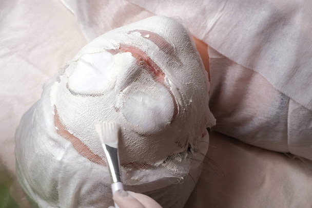Tratamiento de terapia de spa para una mujer musulmana que recibe una máscara en un salón de belleza. El esteticista con guantes blancos trabaja con un cepillo cosmético. El procedimiento de rejuvenecimiento y limpieza de la piel en el centro de cosmetología
. - Foto, Imagen