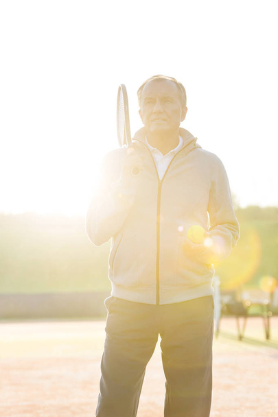 Back lit of senior man standing with tennis racket and ball against clear sky on sunny day - Photo, image