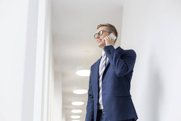 Smiling young businessman talking on smartphone while standing in office corridor - Foto, afbeelding