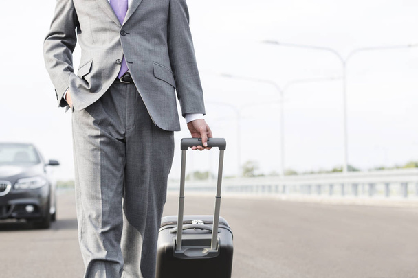 Midsection of businessman walking with suitcase on road against sky - Foto, afbeelding
