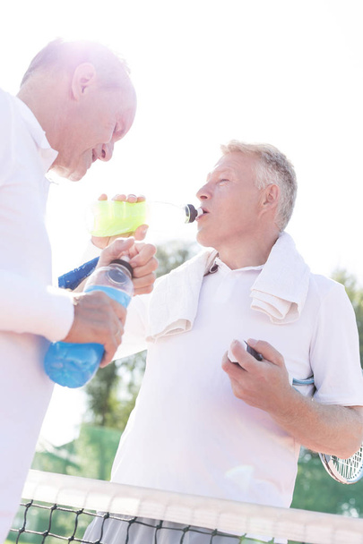 Tiefansicht durstiger Männer, die trinken, während sie bei einem älteren Freund auf dem Tennisplatz vor klarem Himmel stehen - Foto, Bild