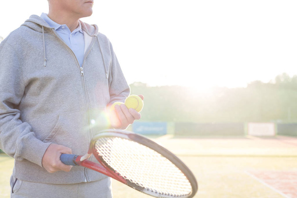 Midsection of mature man holding tennis balls and racket on court against clear sky - Фото, изображение