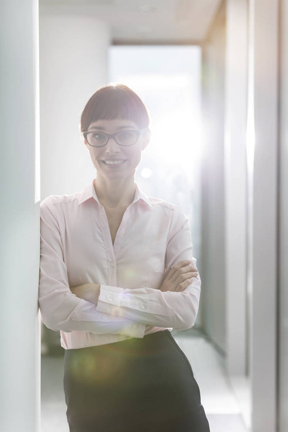 Портрет улыбающейся деловой женщины, стоящей с руками, скрещенными у дверей офиса
 - Фото, изображение