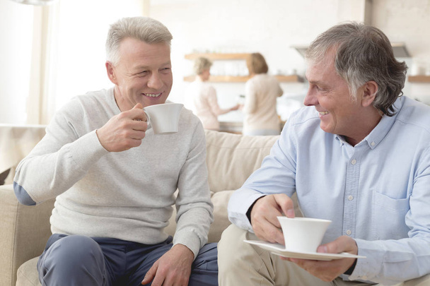 Smiling mature men holding coffee while sitting on sofa at home - Fotografie, Obrázek
