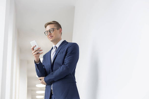 Jeune homme d'affaires souriant utilisant un smartphone tout en restant debout dans le couloir du bureau
 - Photo, image