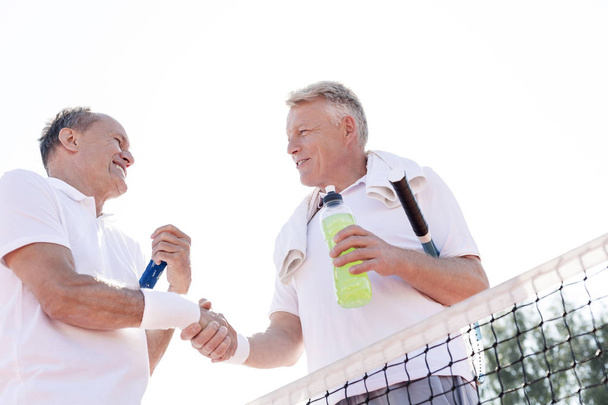 Low angle view of smiling men shaking hands while standing at tennis court against clear sky on sunny day - Photo, Image