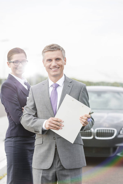 Portrait of smiling insurance agent with clipboard and businesswoman standing against breakdown car - Foto, Bild