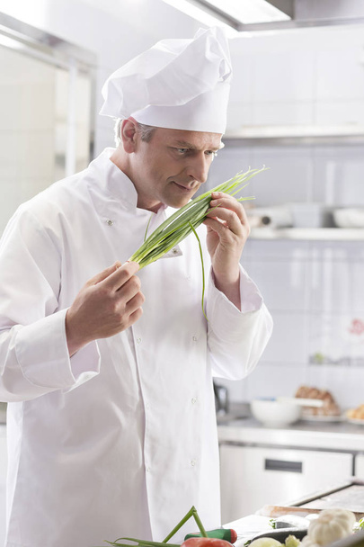 Confident chef smelling fresh scallion bunch while standing at restaurant kitchen - Φωτογραφία, εικόνα