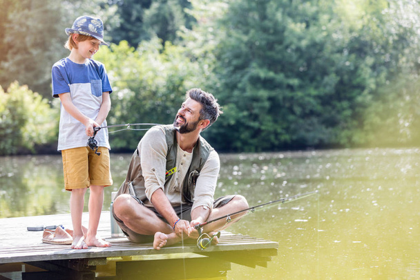 Усміхнений батько дивиться на сина, що стоїть на пірсі під час риболовлі в озері
 - Фото, зображення