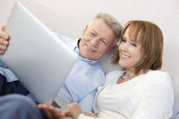 Smiling mature couple using laptop in bedroom at home - Fotó, kép