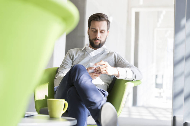 Confident businessman using smart phone in coffee break at office - Фото, изображение
