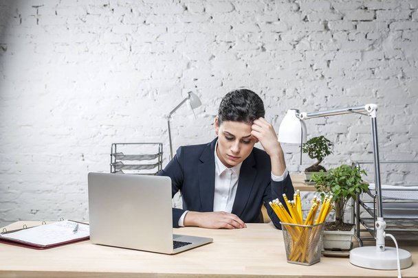 Bored young businesswoman sitting with laptop at desk against brick wall in office - Foto, afbeelding