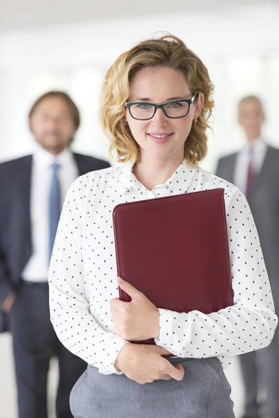 Portrait of confident businesswoman with file standing against colleagues at office - Fotoğraf, Görsel