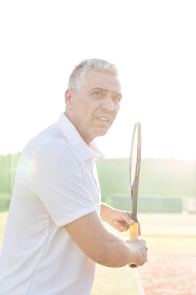Zijaanzicht van zelfverzekerde volwassen man swingen tennisracket op rechter tegen heldere hemel - Foto, afbeelding