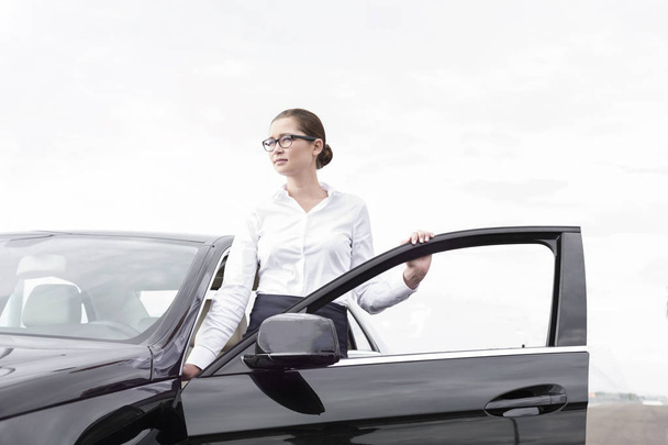 Young businesswoman standing by open door of car against sky - Foto, afbeelding