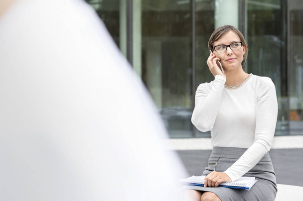 Confident businesswoman talking on smartphone while sitting against office building - Photo, Image