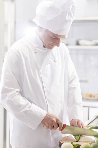 Confident chef cutting cucumber on board in restaurant kitchen - Zdjęcie, obraz