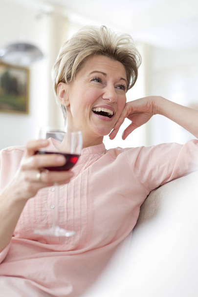 Cheerful mature woman holding wineglass while looking away at home - Foto, Imagem