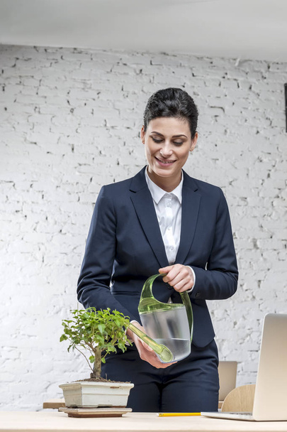 Smiling young businesswoman watering plant on desk against brick wall at office - Foto, Bild