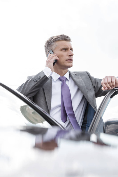 businessman talking on mobile phone while standing outside car against sky - Photo, Image