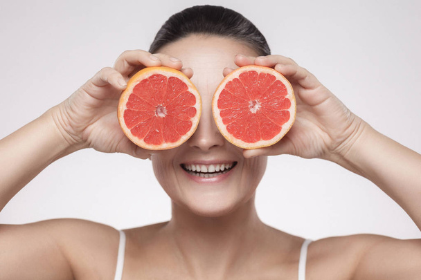 Portrait of middle aged woman holding fresh slices of grapefruit on grey background - Photo, Image
