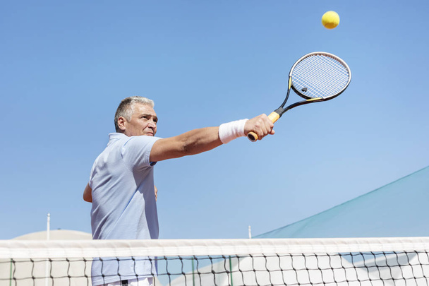 Low angle view of confident mature man hitting tennis ball with racket on court against clear blue sky - Photo, image