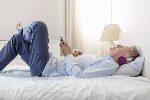Side view of man listening music while lying on bed at home - Foto, afbeelding