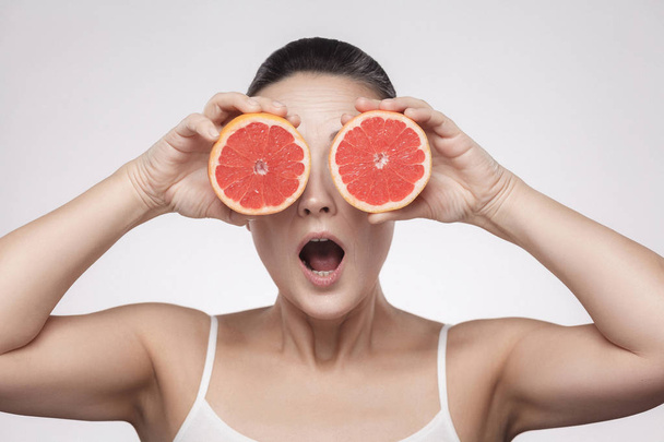 Portrait of middle aged woman holding fresh slices of grapefruit on grey background - Zdjęcie, obraz