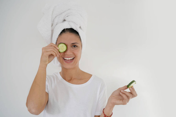 Vibrant brunette with towel turban and cucumber on eye on white background - Foto, Imagem