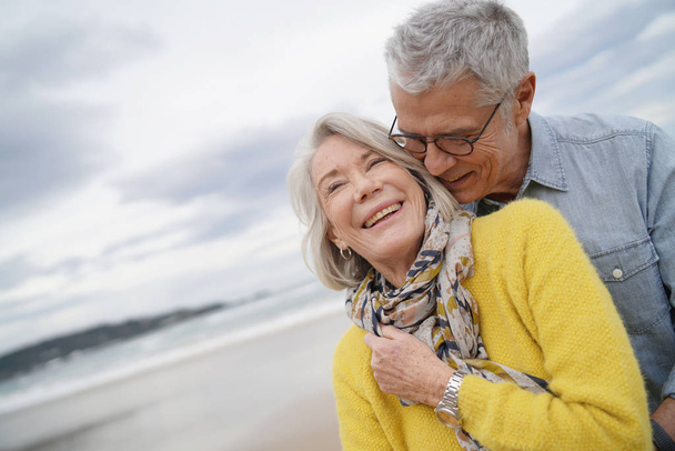 Retrato de atractiva pareja de ancianos vibrantes abrazándose en la playa en otoño
                               - Foto, imagen